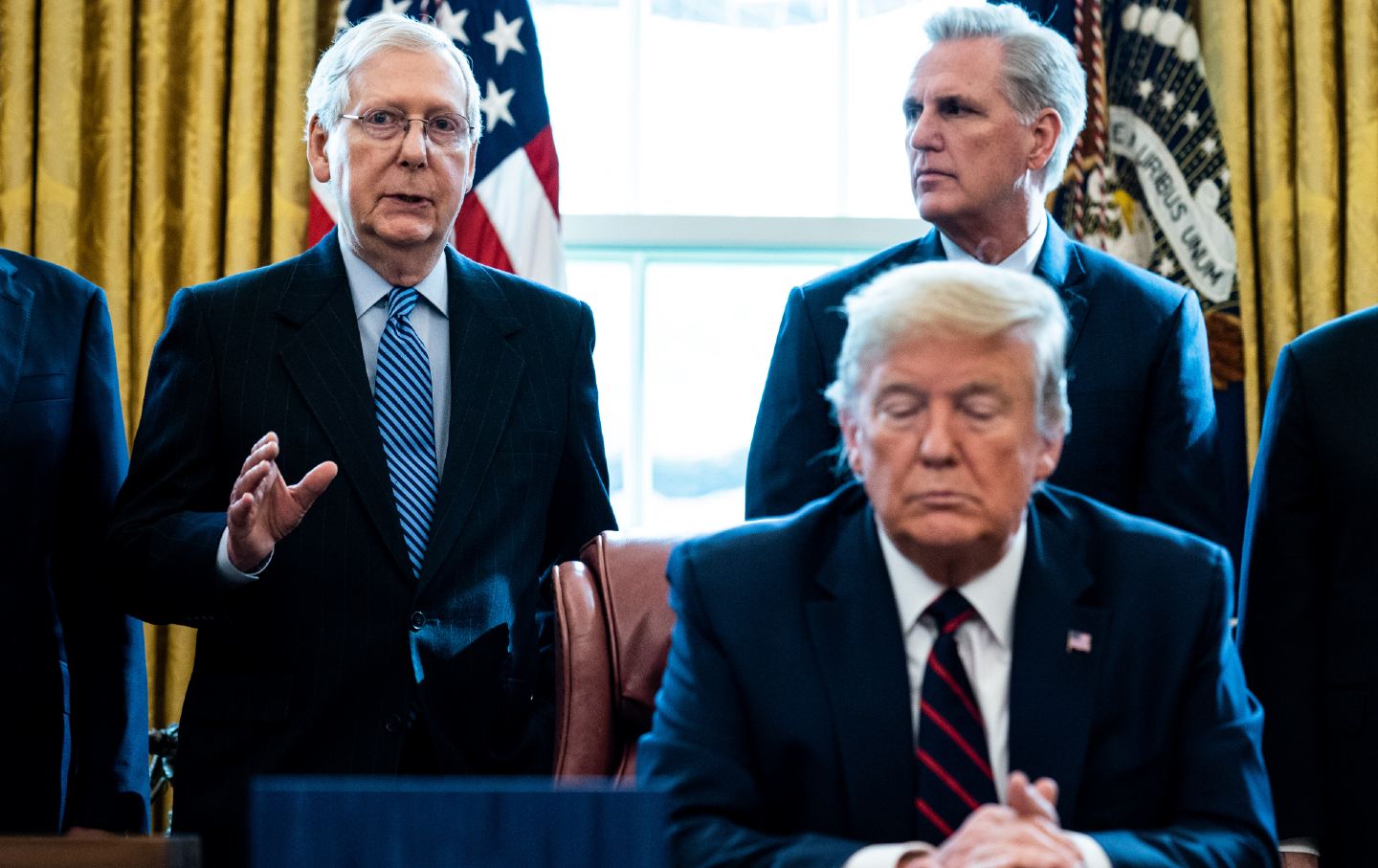 Donald Trump sits at a desk with Mitch McConnell and Kevin McCarthy standing behind him