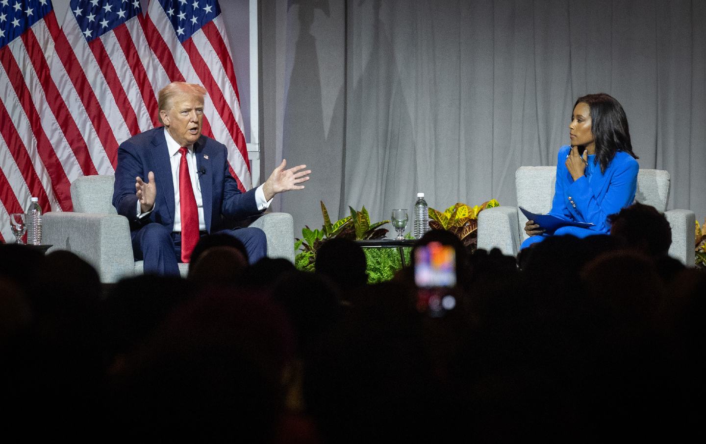 Donald Trump sitting on stage at a panel in front of American flag