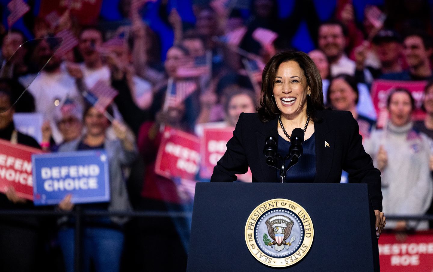 Vice President Kamala Harris beams at a podium in front of a crowd holding signs reading 