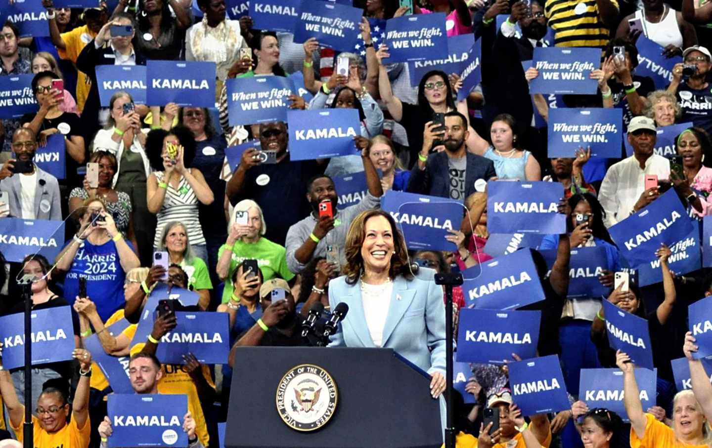 Vice President Kamala Harris smiles at a podium, in front of many people holding signs reading 