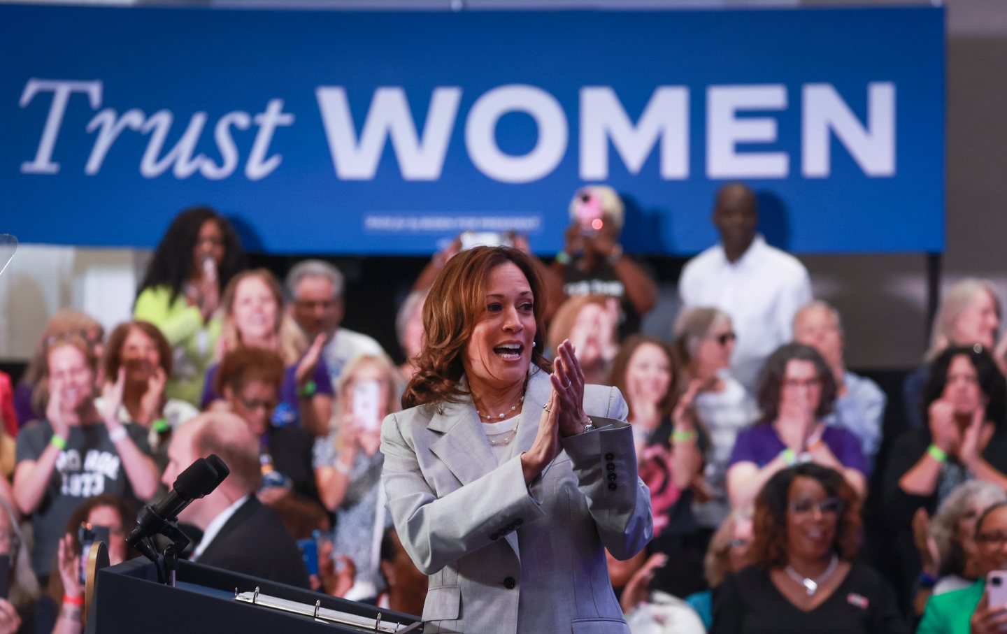 Vice-President Kamala Harris claps and smiles at a podium in front of a banner reading 