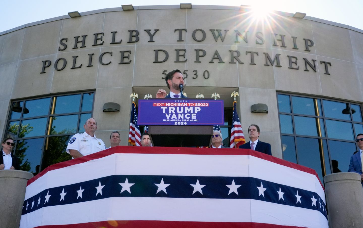 Sen. JD Vance at a podium above bunting, in front of the Shelby Township Police Department, flanked by police officers.