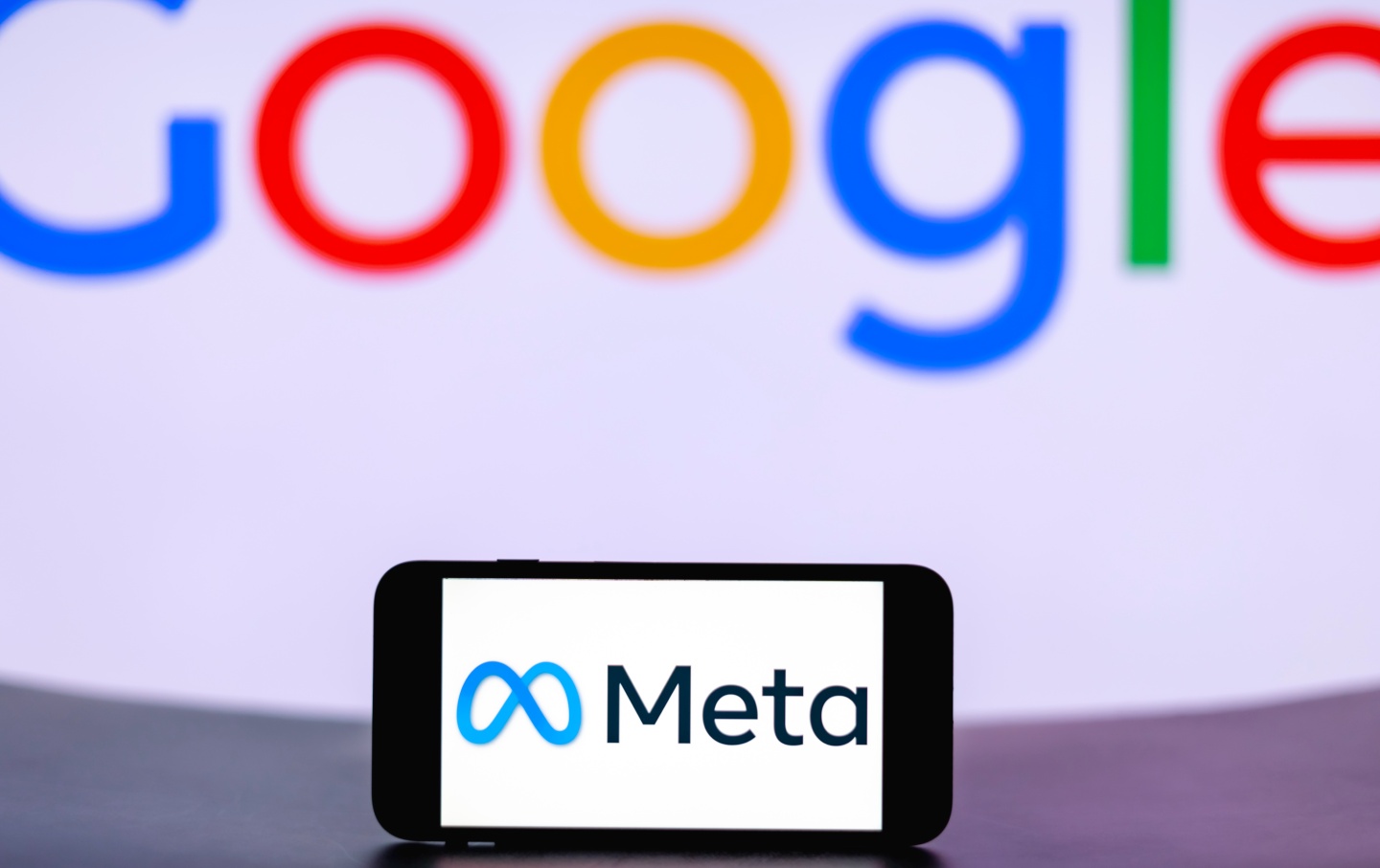 A phone screen shows the logo of Meta, with the logo of Google looming in the background.