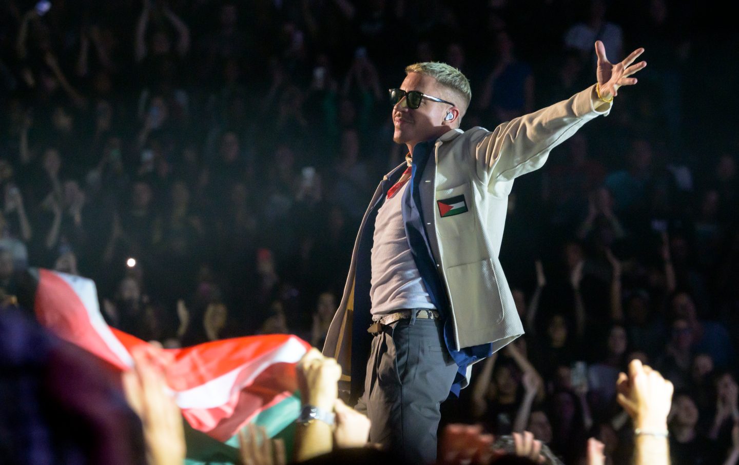 Macklemore, wearing sunglasses and a jacket with a Palestinian flag, gestures at a crowd in a concert.