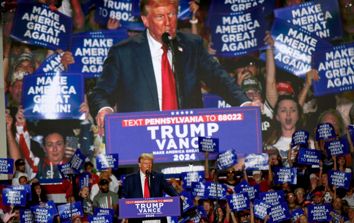 Former President Donald Trump on stage in front of a large crowd, and also in front of a Jumbotron image of him at the podium.