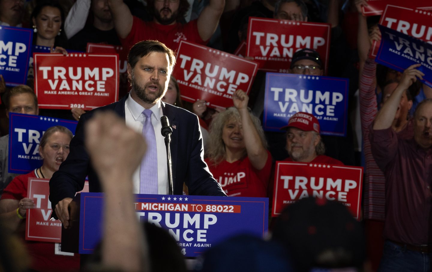 Sen. J.D. Vance onstage at a rally, surrounded by Trump/Vance signs.