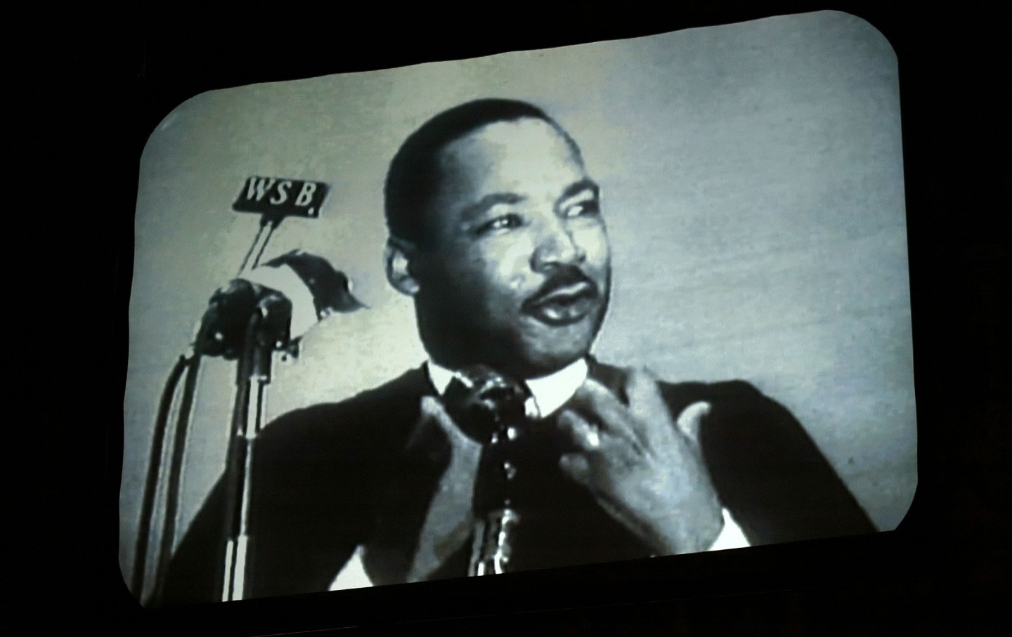 Foto hitam-putih Pendeta Dr. Martin Luther King Jr., di TV, berbicara di depan mikrofon.