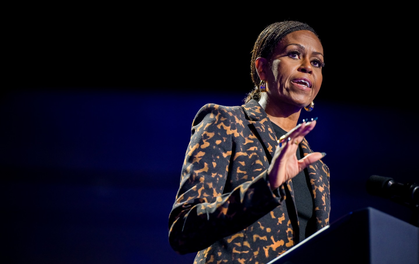 Former First Lady Michelle Obama at a podium, speaking and holding up her hand in a gesture.