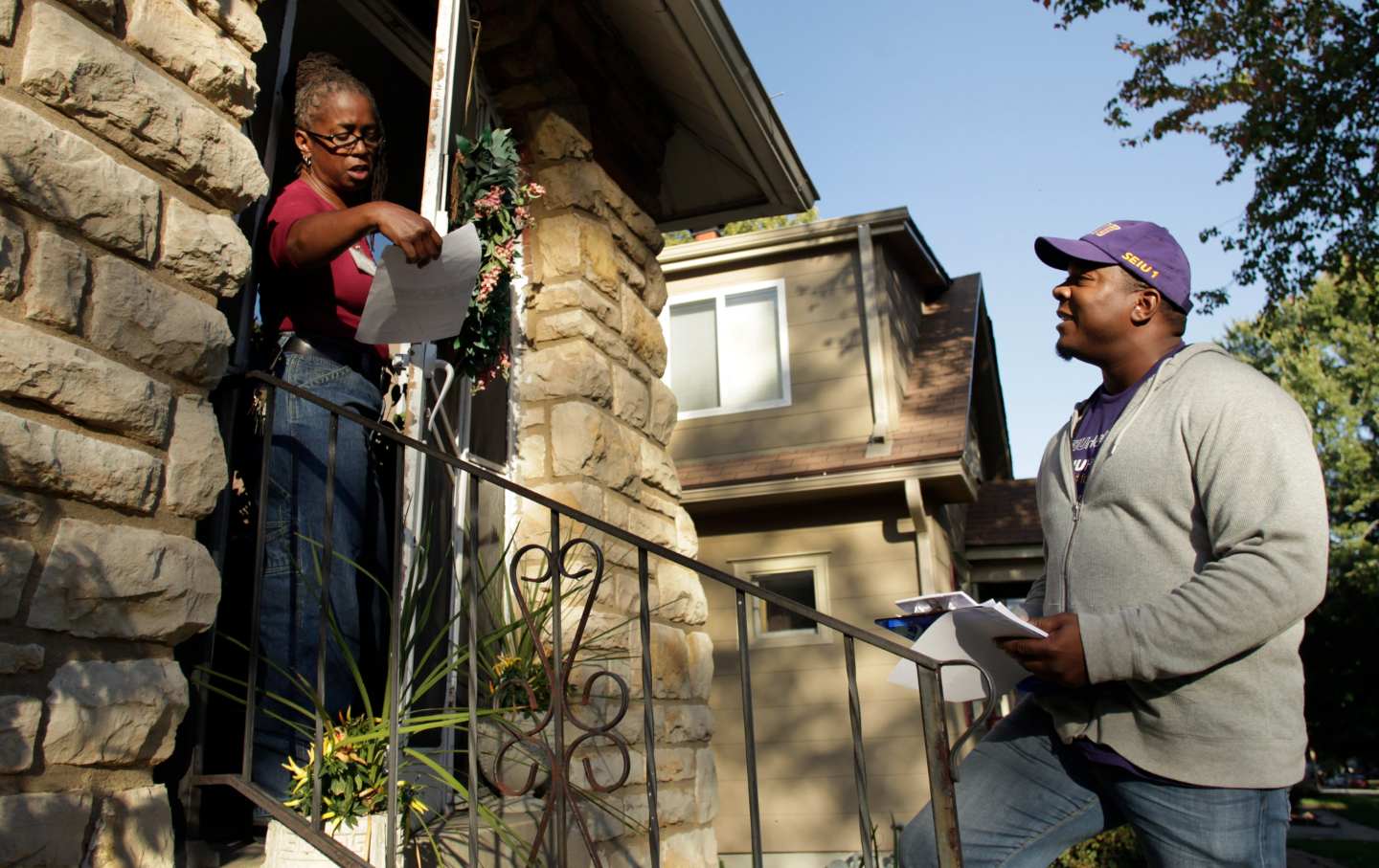 A canvasser stands on a voter’s stoop having handed over campaign literature. The voter is reading the pamphlet.