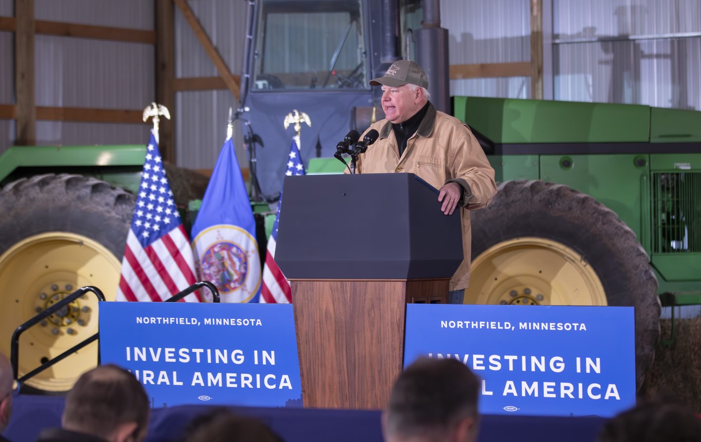 Gubernur Minnesota Tim Walz berbicara di depan podium, dengan traktor dan bendera Amerika di belakangnya