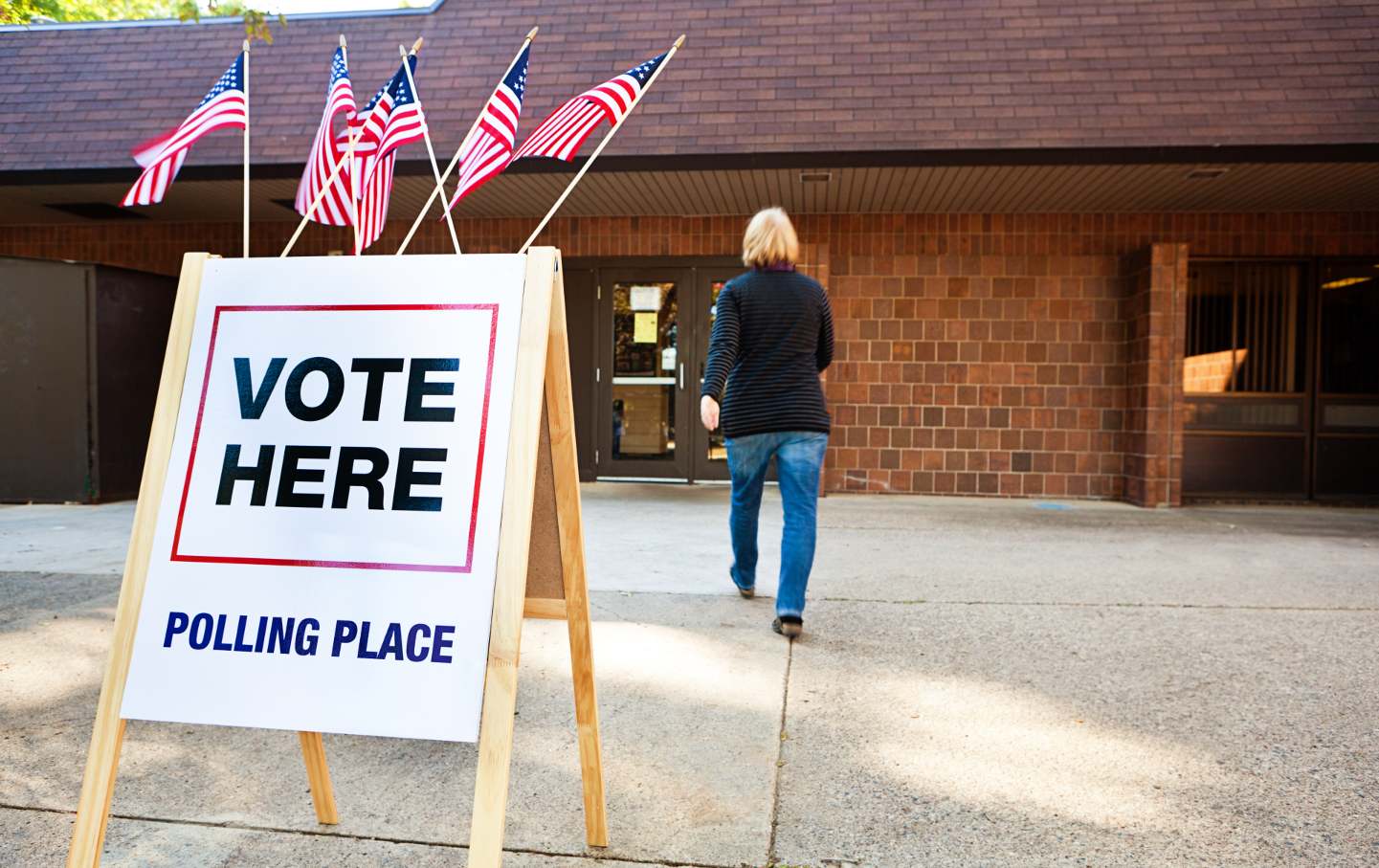 In the foreground, a "Vote Here" sign with small American flags flying above it; in the background, a white woman walks toward a polling place.