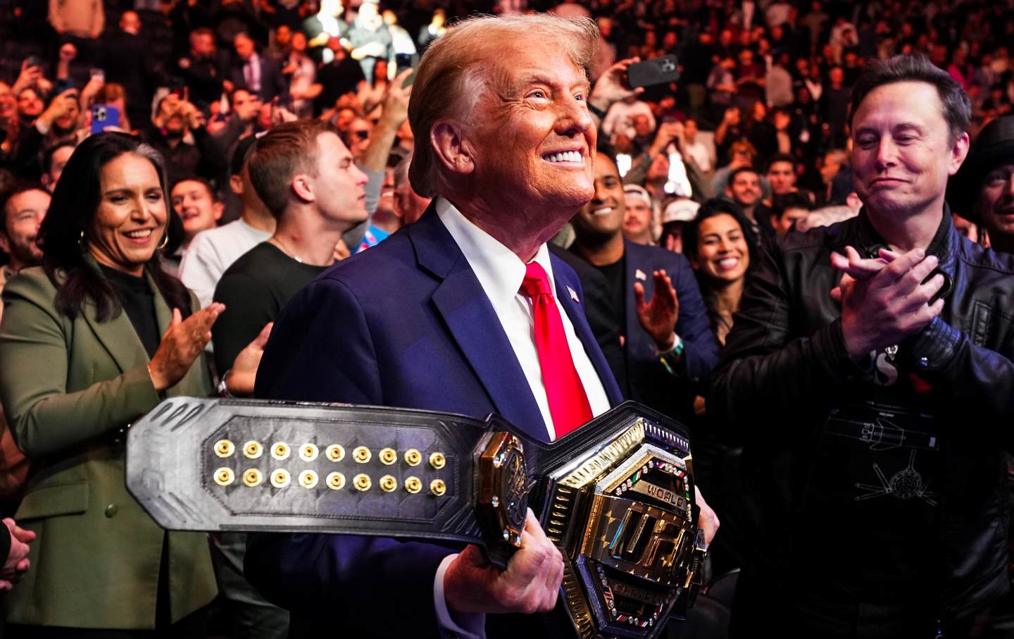 A grinning Trump holds up the UFC belt. Tulsi Gabbard and Elon Musk stand in crowd behind him, clapping.