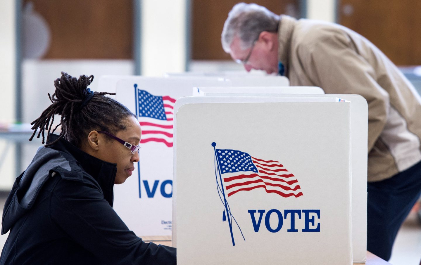 In the foreground, a Black woman at a voting booth; in the background, a white man at a voting booth.