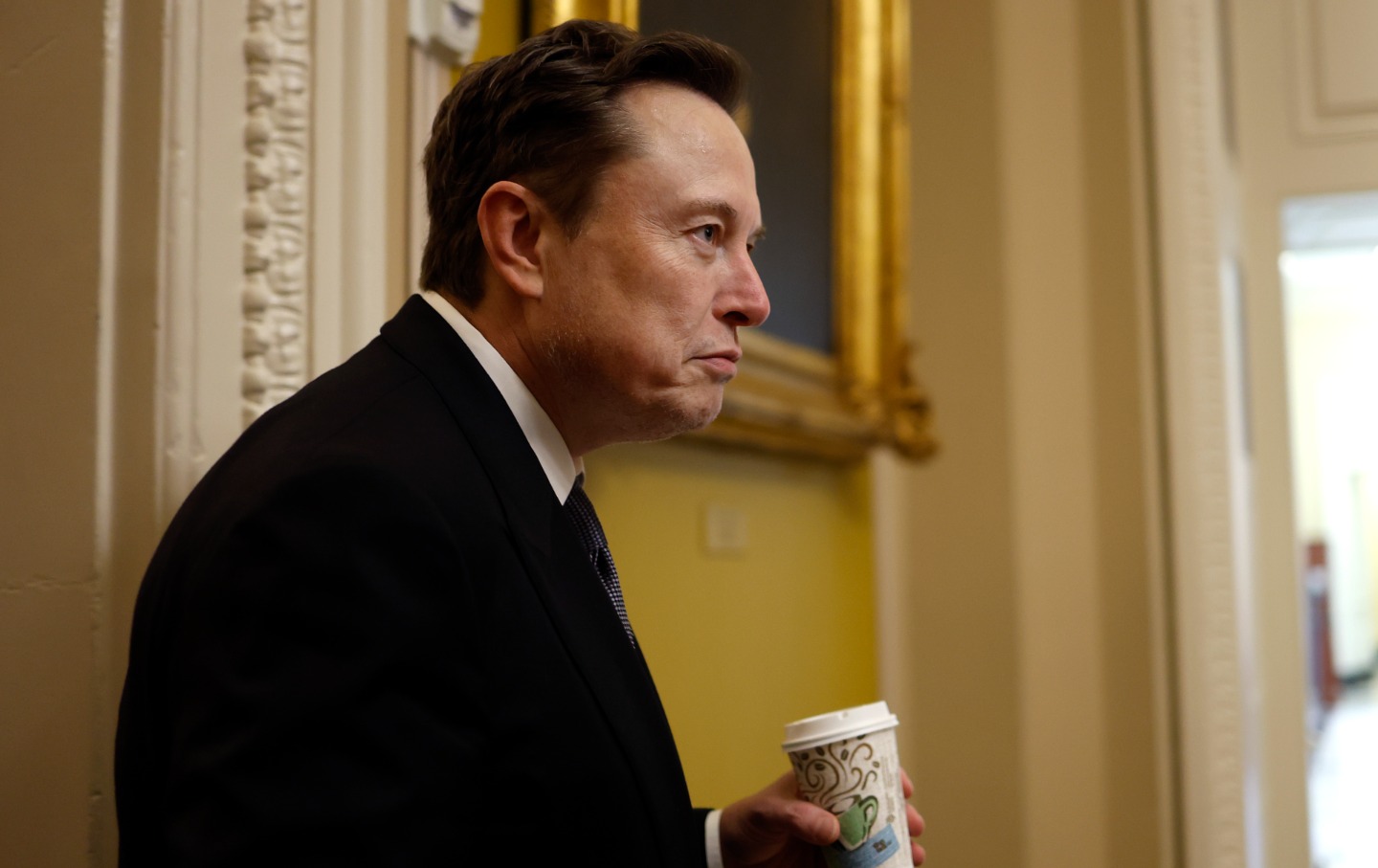 Elon Musk inside the Capitol Building, holding a cup of coffee and frowning.