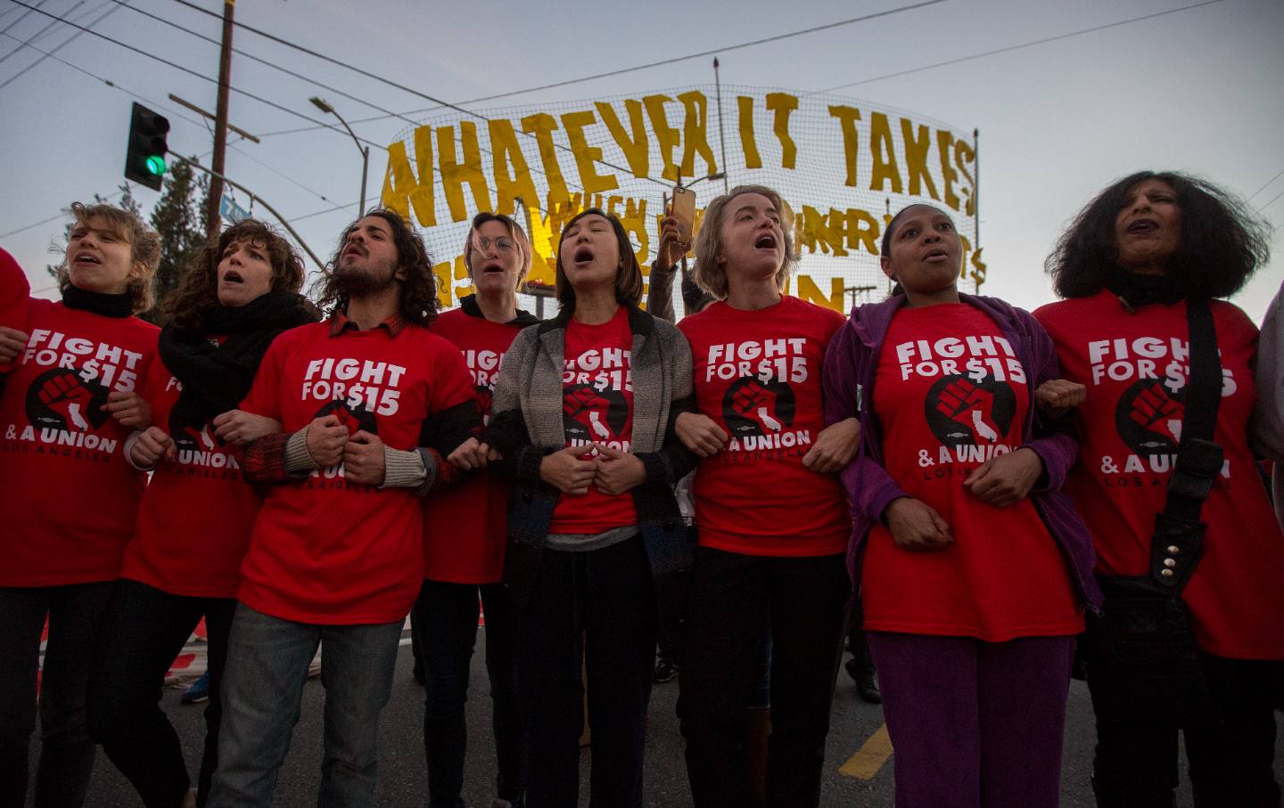 Workers in red “Fight for $15” T-shirts standing next to each other with locked arms, in front of a banner reading “Whatever It Takes.”