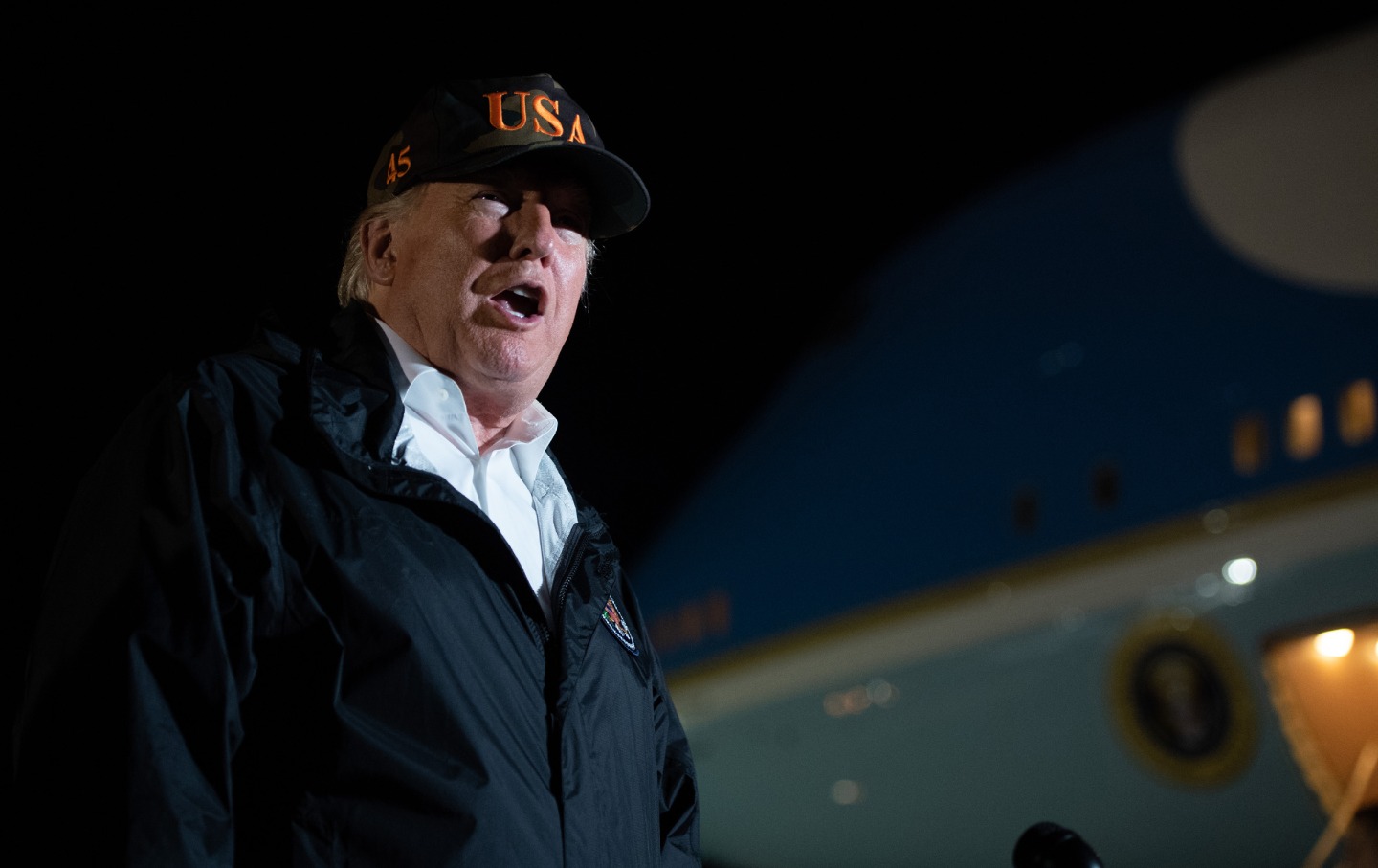 Donald Trump at night, in front of Air Force One, wearing a USA baseball cap.