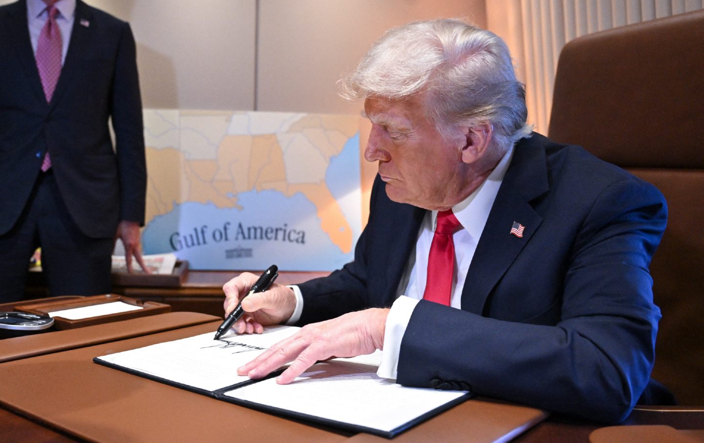 Donald Trump sits at a desk, signing a document. Behind him, a map reads 