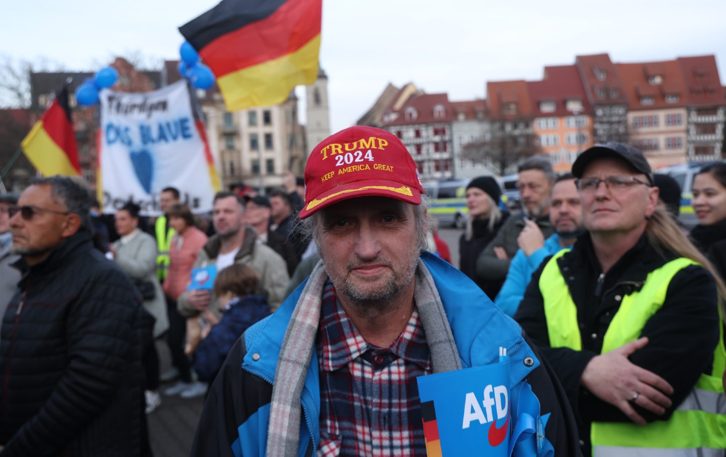 A man standing in a crowd. He has a hat that reads "Trump 2024 Keep America Great" and is carrying an AfD flag.