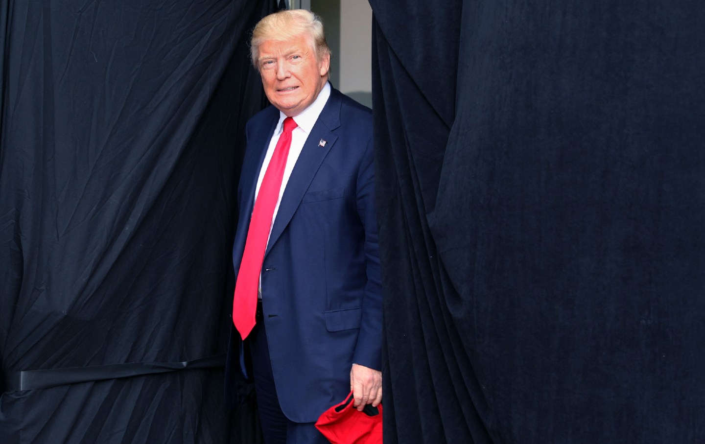 Donald Trump stepping out onto a stage while holding a red MAGA hat.