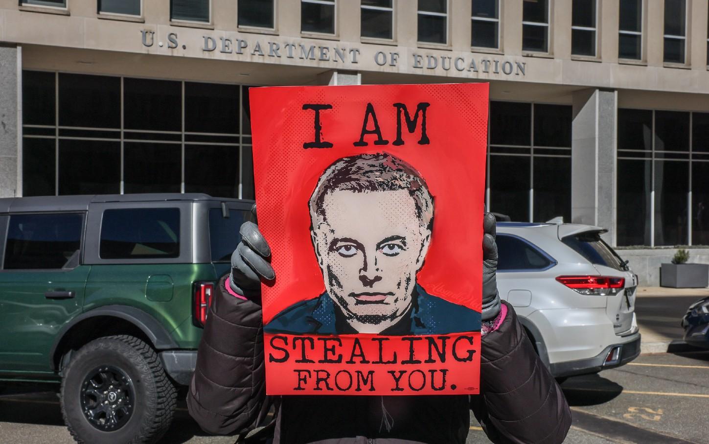 A protester holds a poster with an image of Elon Musk that reads “I Am Stealing From You” at the US Department of Education in Washington, DC, on February 14, 2025.