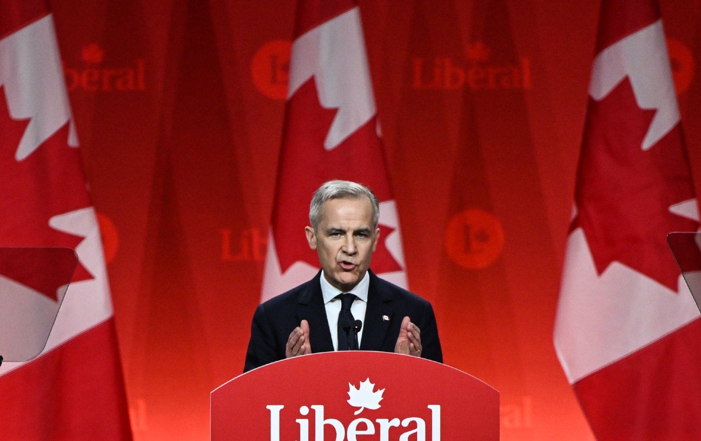 A man stands at a podium. Behind him, there are three Canadian flags and a background that reads 