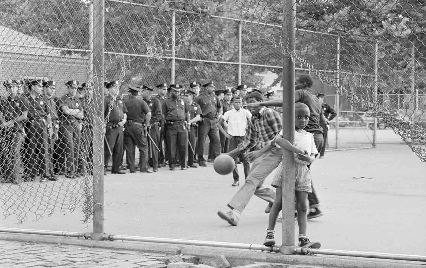 A young boy peers out from a hole in a fence as his friends play basketball in a court where police officers are gathering for a patrol in East New York, 1966.